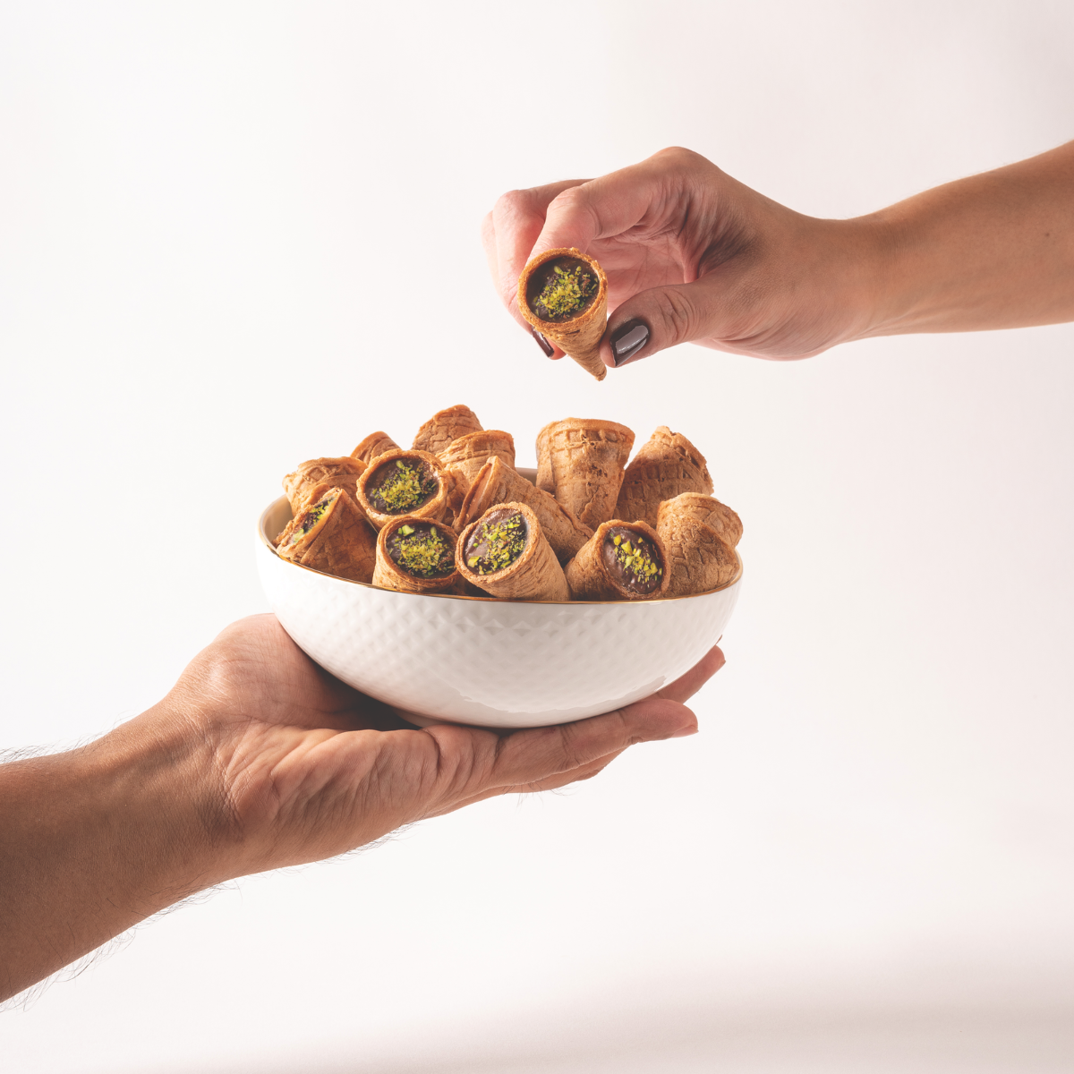 Two hands holding a white bowl filled with Leonessa Dubai Chocolate Cones, against a white background